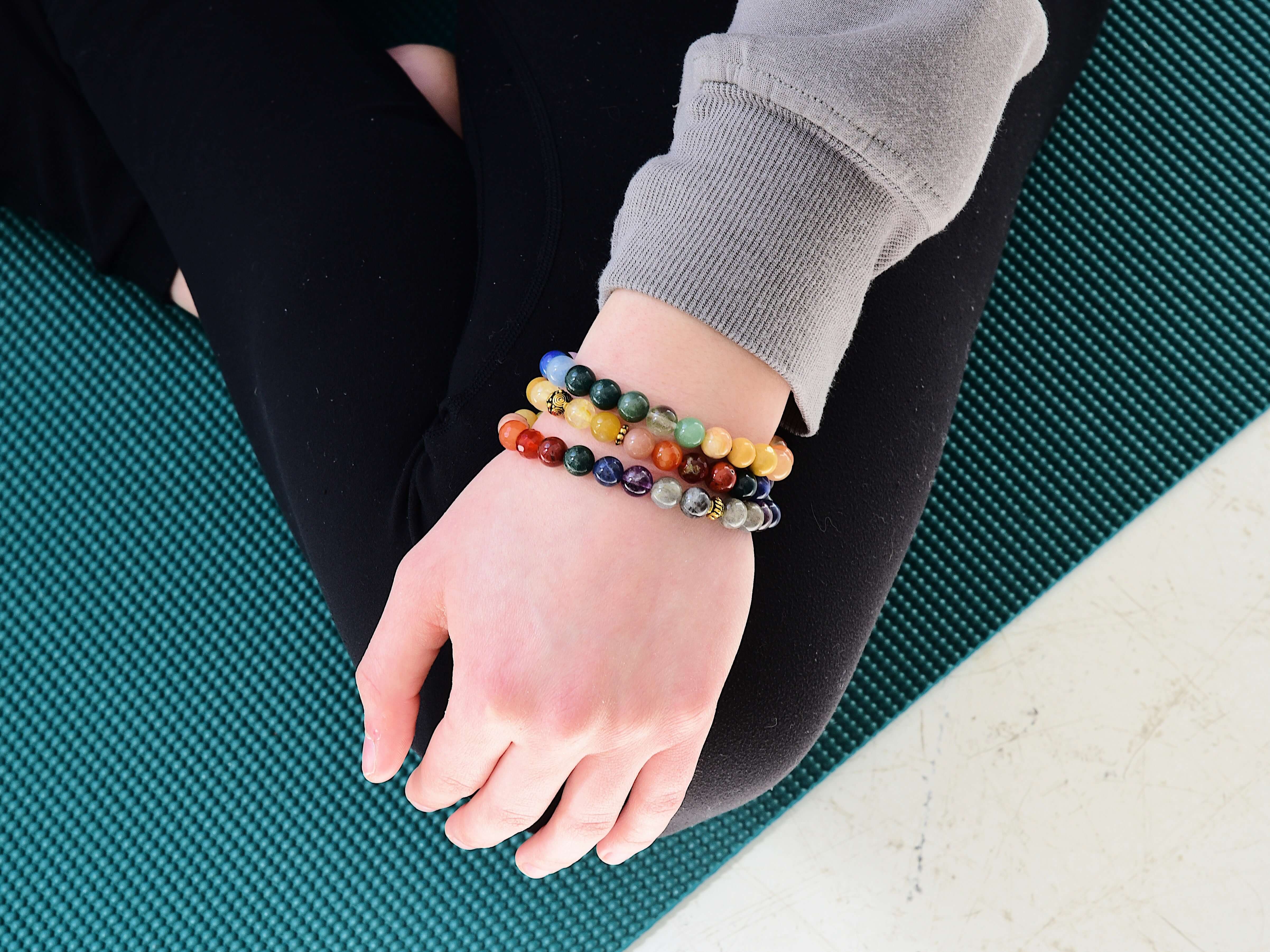 Close up of a Rainbow Love Bracelet and two Divine Feminine bracelets on a wrist of a person sitting on a yoga mat.