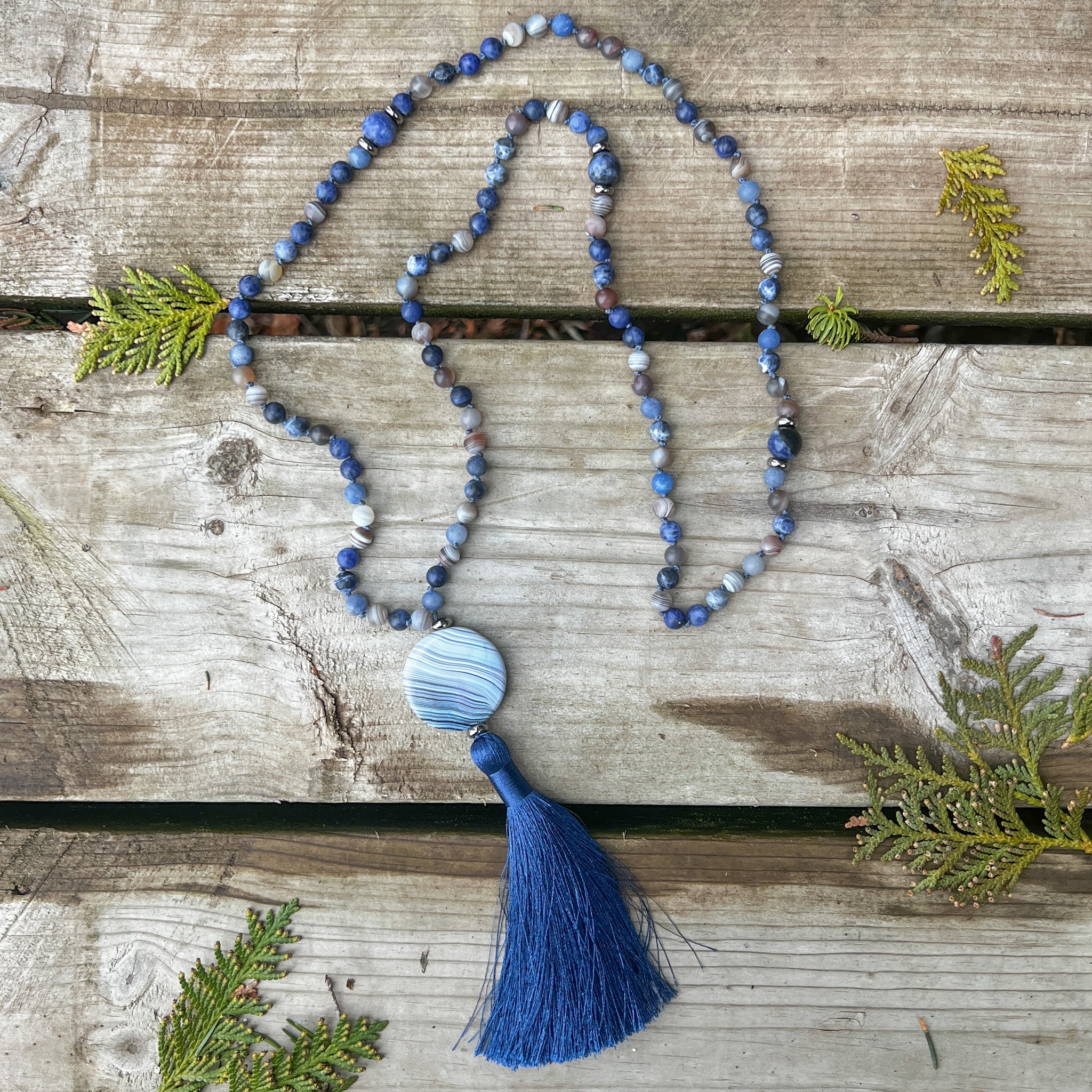 Ocean Current Mala Necklace with Blue Aventurine, Sodalite, Botswana Agate, blue agate focal stone, and navy silk tassel on weathered wood.