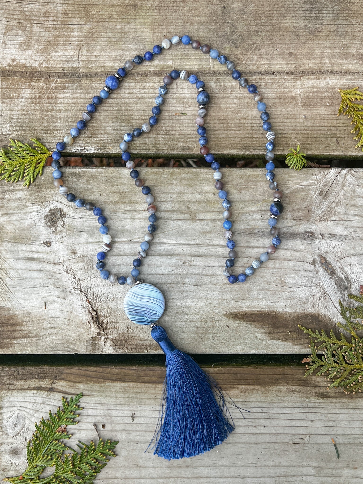 Handmade Ocean Current Mala Necklace featuring 108 gemstones and a blue agate focal pendant displayed on rustic wooden boards.