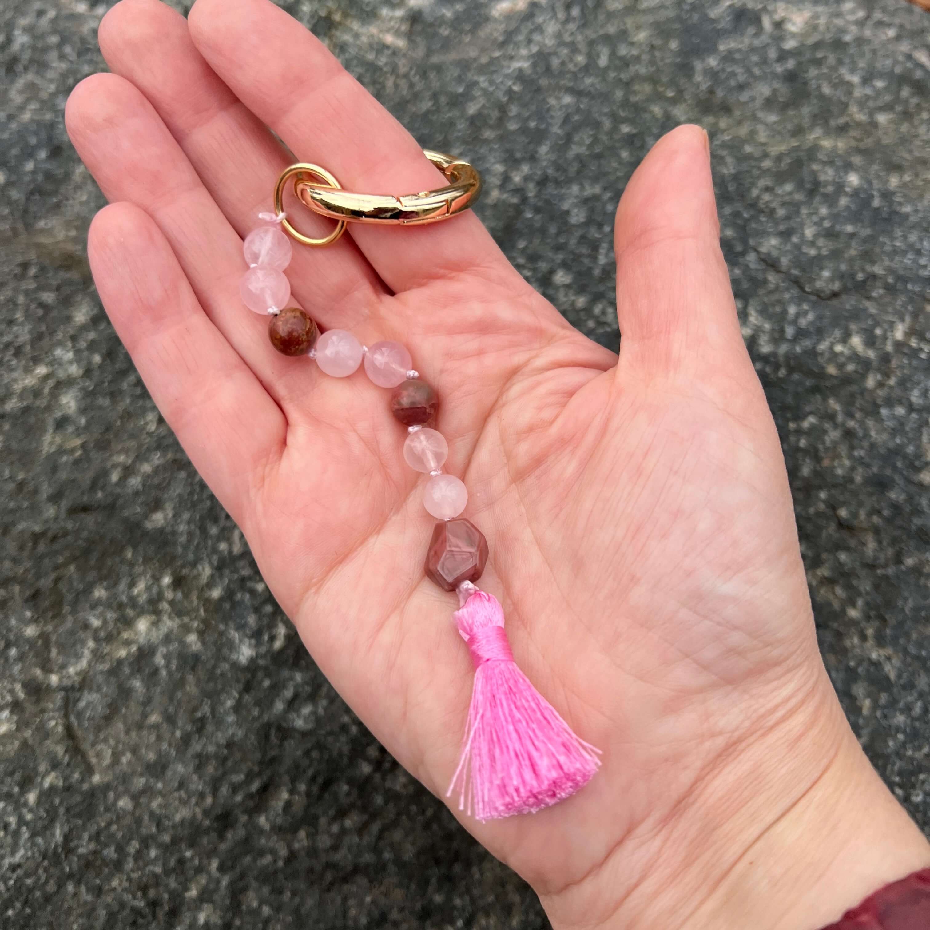 Hand holding a small beaded keychain with a pink tassel against a stone background