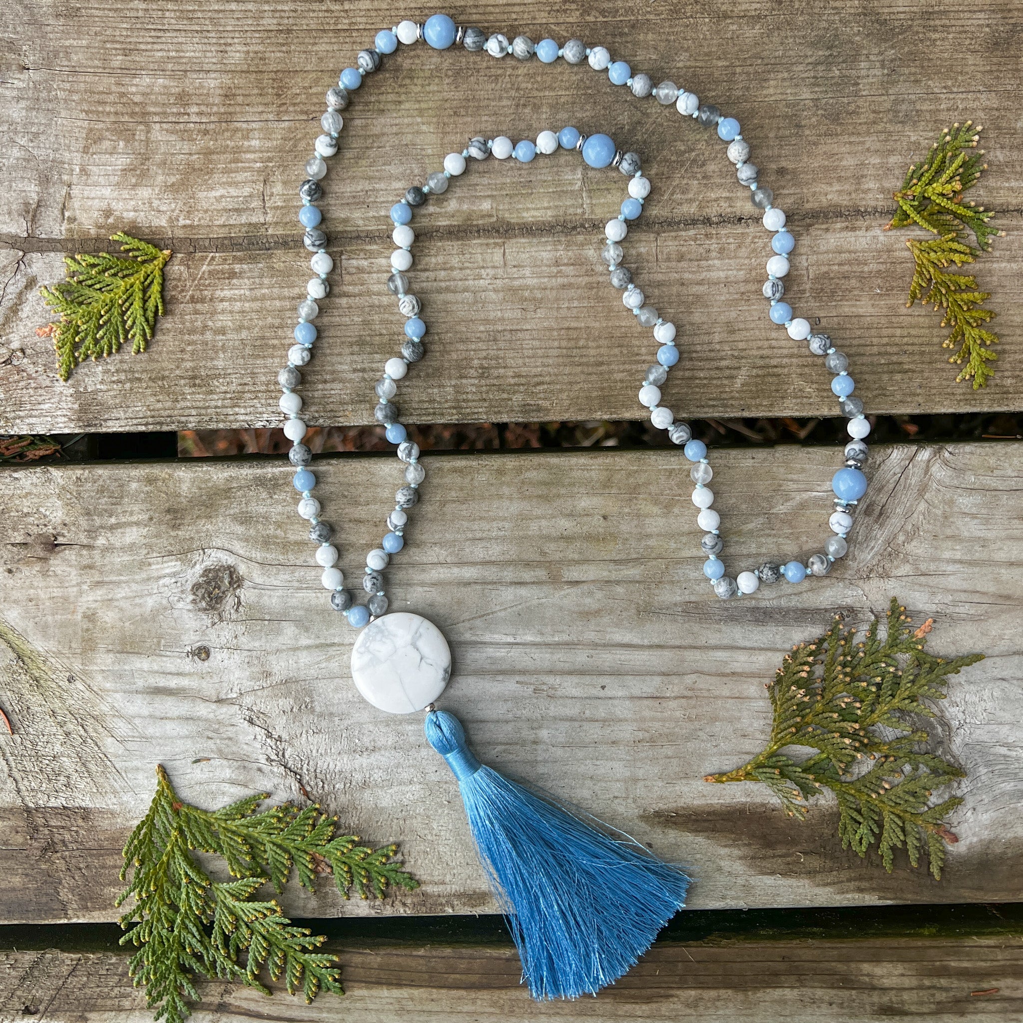 Open Sky Mala Necklace with Howlite, Angelite, Silver Crazy Lace Agate, and a sky blue tassel displayed on weathered wood with cedar sprigs.