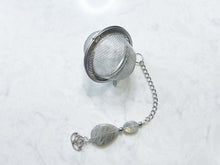 Load image into Gallery viewer, Labradorite Tea Strainer This tea strainer is made with a stainless steel tea ball, Labradorite and Hematite stones with a lotus flower charm. These stones bring the user protection and encourages spiritual transformation.
